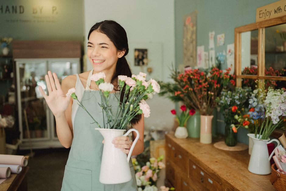 A young woman with long dark hair, smiling warmly and waving, stands inside a flower shop holding a white ceramic pitcher filled with fresh pink and white carnations. She wears a light green apron ove