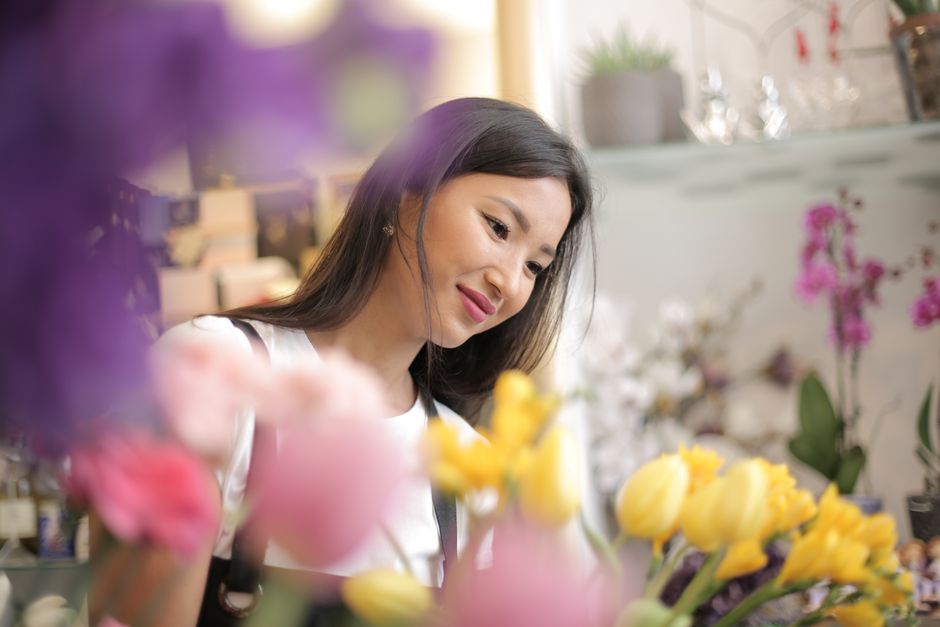 A woman with long dark hair and a gentle smile is inspecting a bouquet of fresh yellow tulips in a floral shop. She is wearing a white top and standing in front of shelves filled with various flowers
