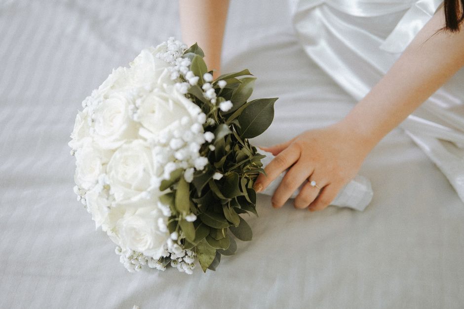 A close-up of a bridal bouquet consisting of white roses, accented with small white filler flowers and lush green foliage, held by a bride dressed in white satin fabric. The bouquet is arranged in a r