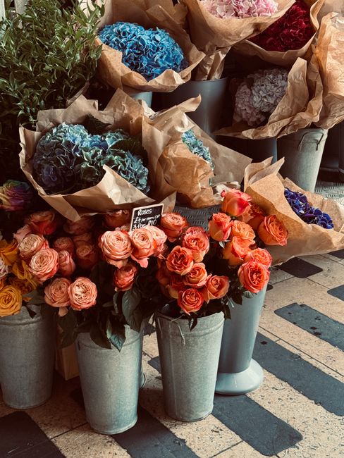 A collection of fresh flower bouquets arranged in tall, gray cylindrical pots at a flower shop display. The bouquets feature pink roses with tightly packed petals, some with slightly open blossoms, an