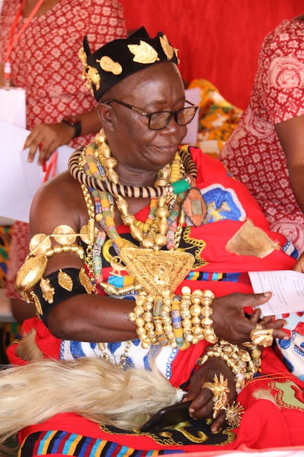 A woman dressed in traditional Maasai attire, wearing a vibrant red shúkà and adorned with elaborate gold and beaded jewelry, including necklaces, bracelets, and earrings. She is seated indoors on a